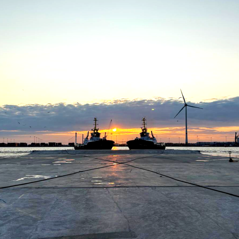 Offshore marine construction site at sunset with vessels and a wind turbine on the horizon.